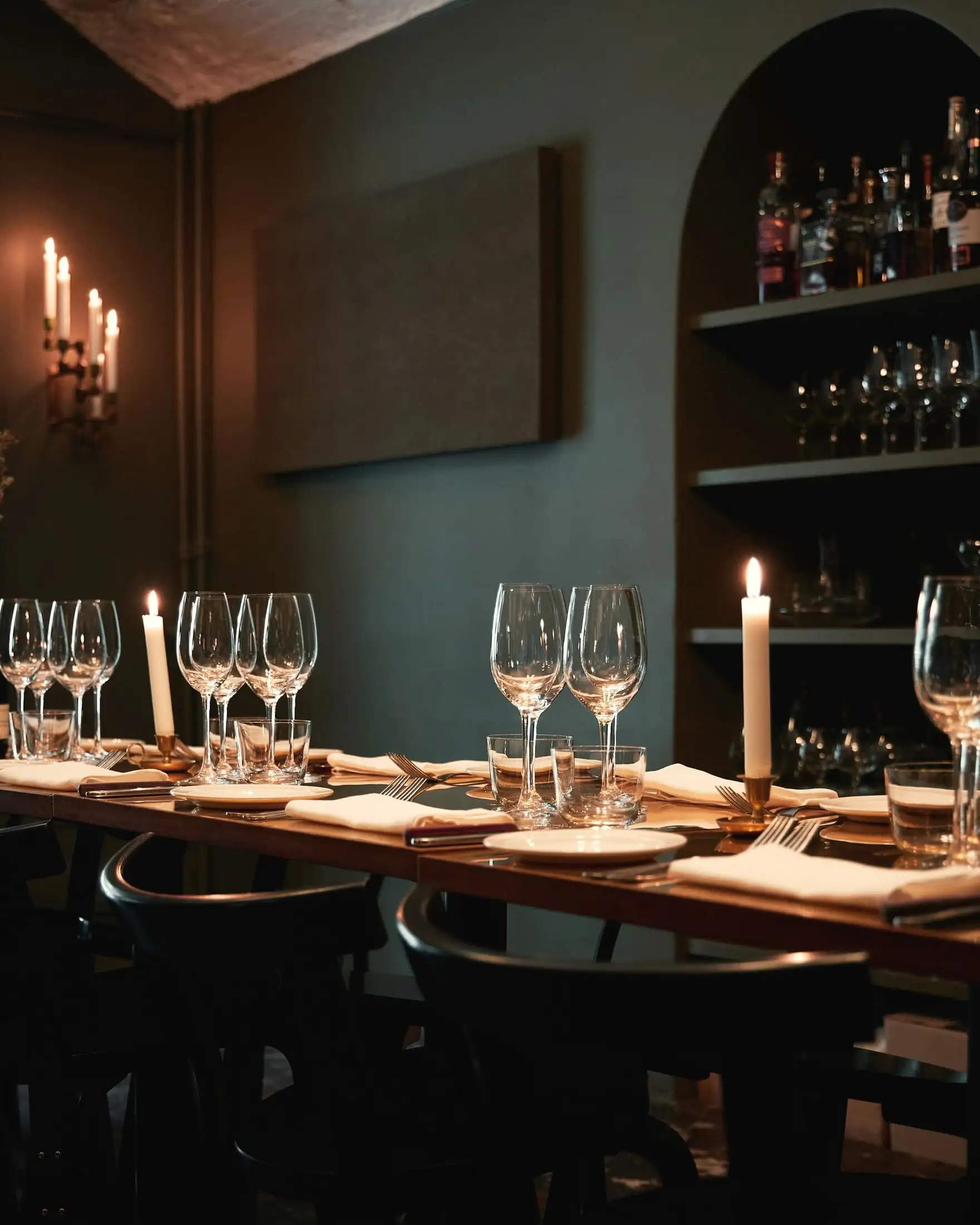 A long wooden table laid with wine glasses and candles in a dark green room with shelves of bottles.