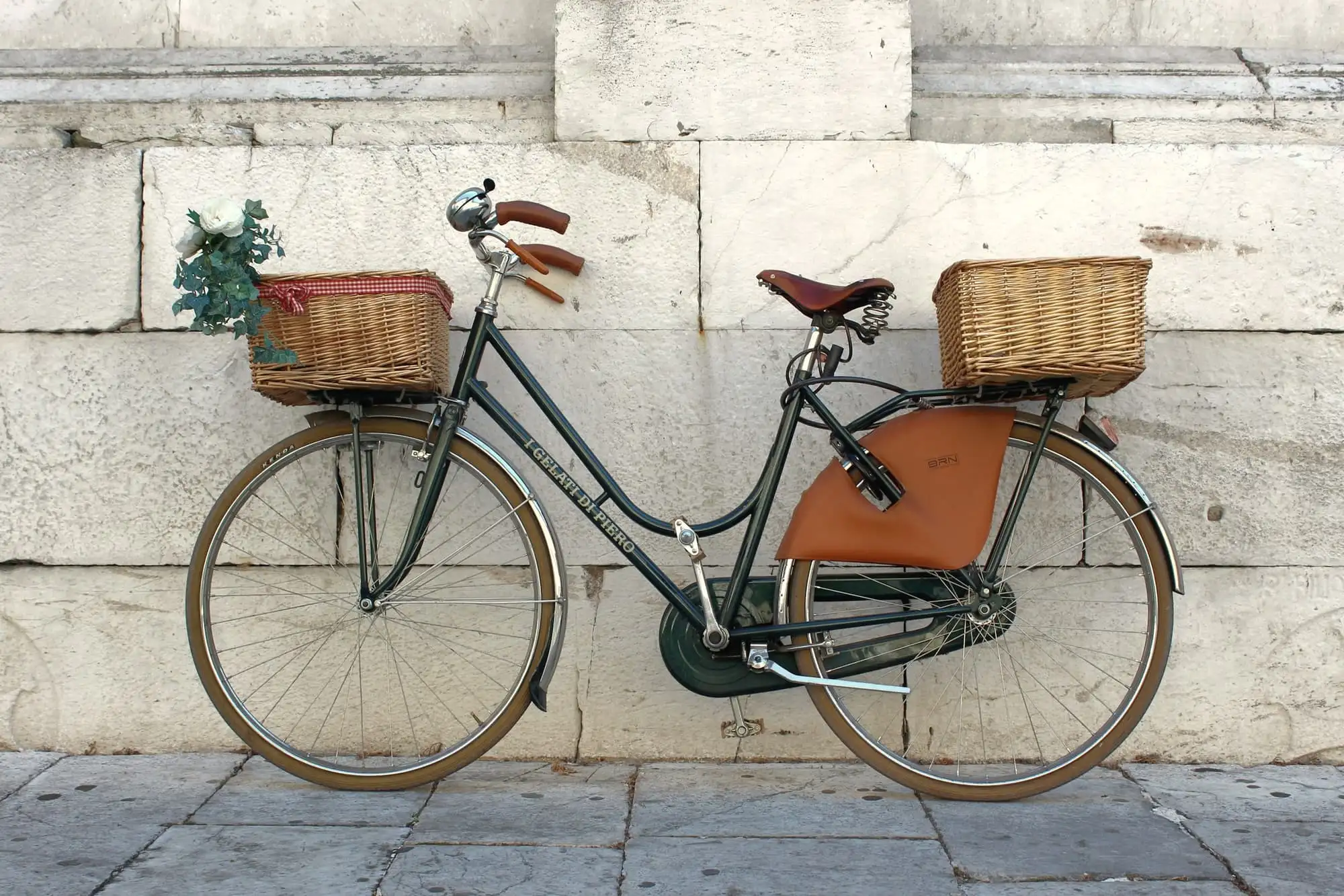 A dark green city bicycle with a wicker basket and a tan leather mudguard, leaning on a pale stone wall.