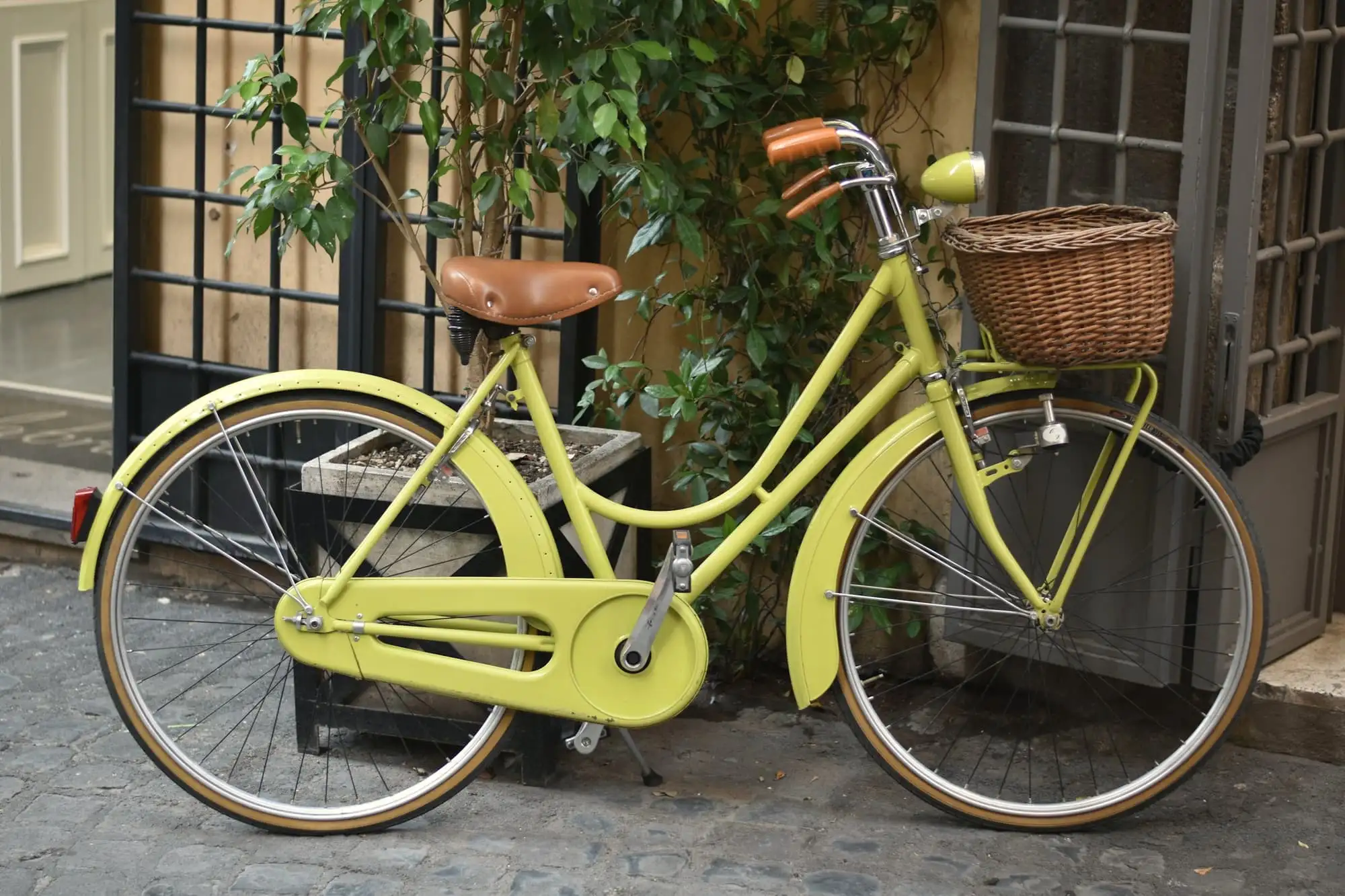 A pale yellow bicycle with a wicker basket, parked on a cobbled lane beside a doorway and a climbing plant.