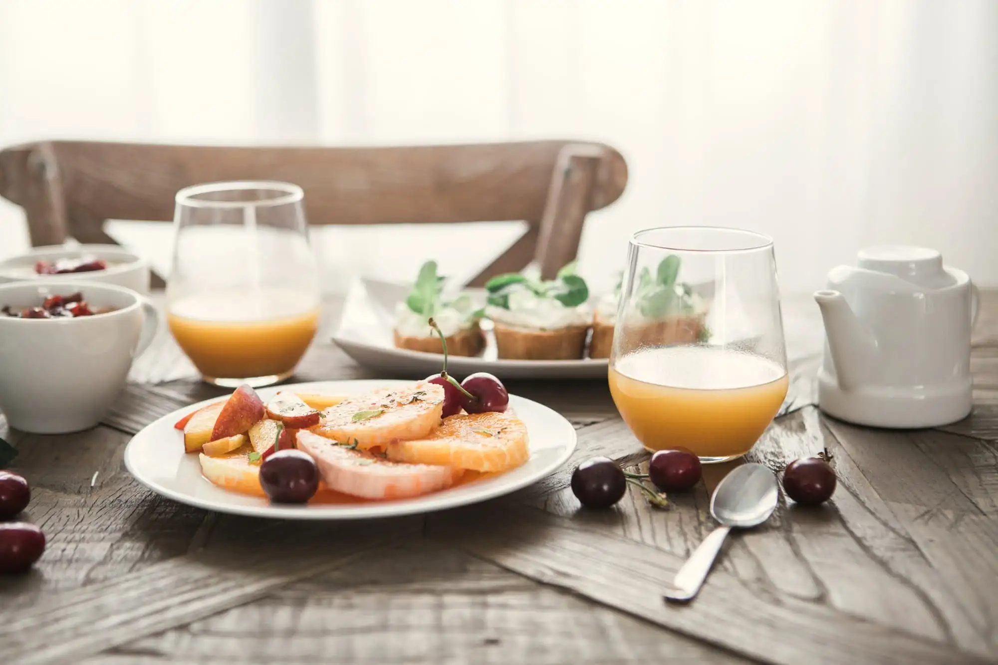 A breakfast table with a plate of sliced orange and cherries, two glasses of juice and a white teapot.