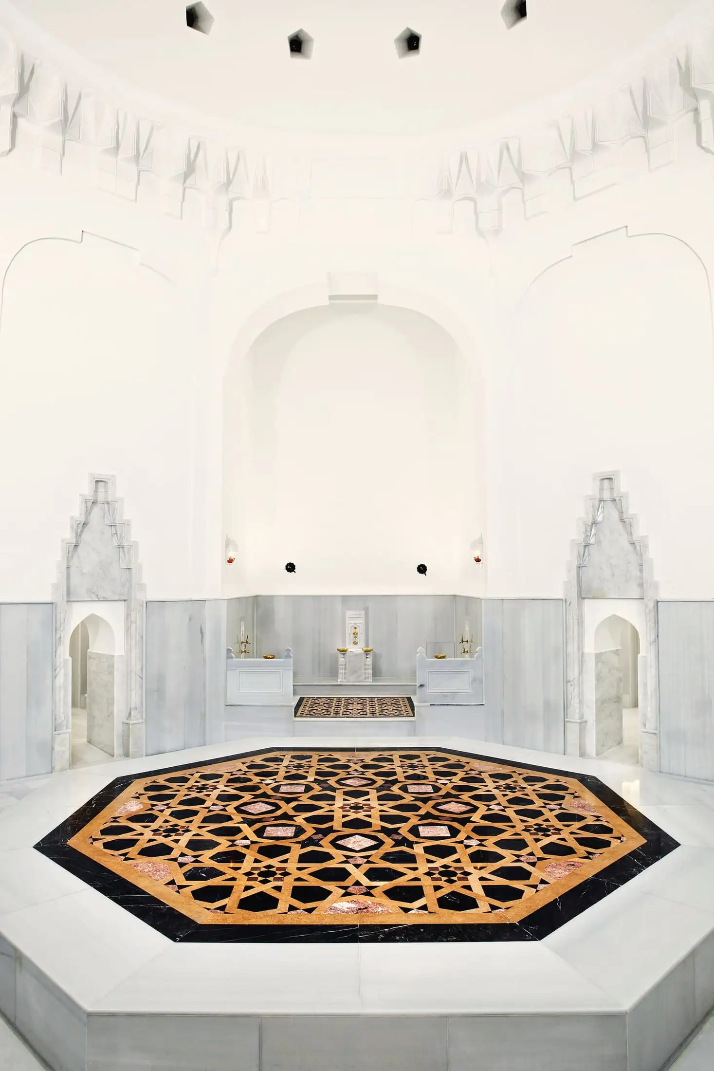 A white marble hammam with two carved arched niches, a basin between them and a patterned stone floor.
