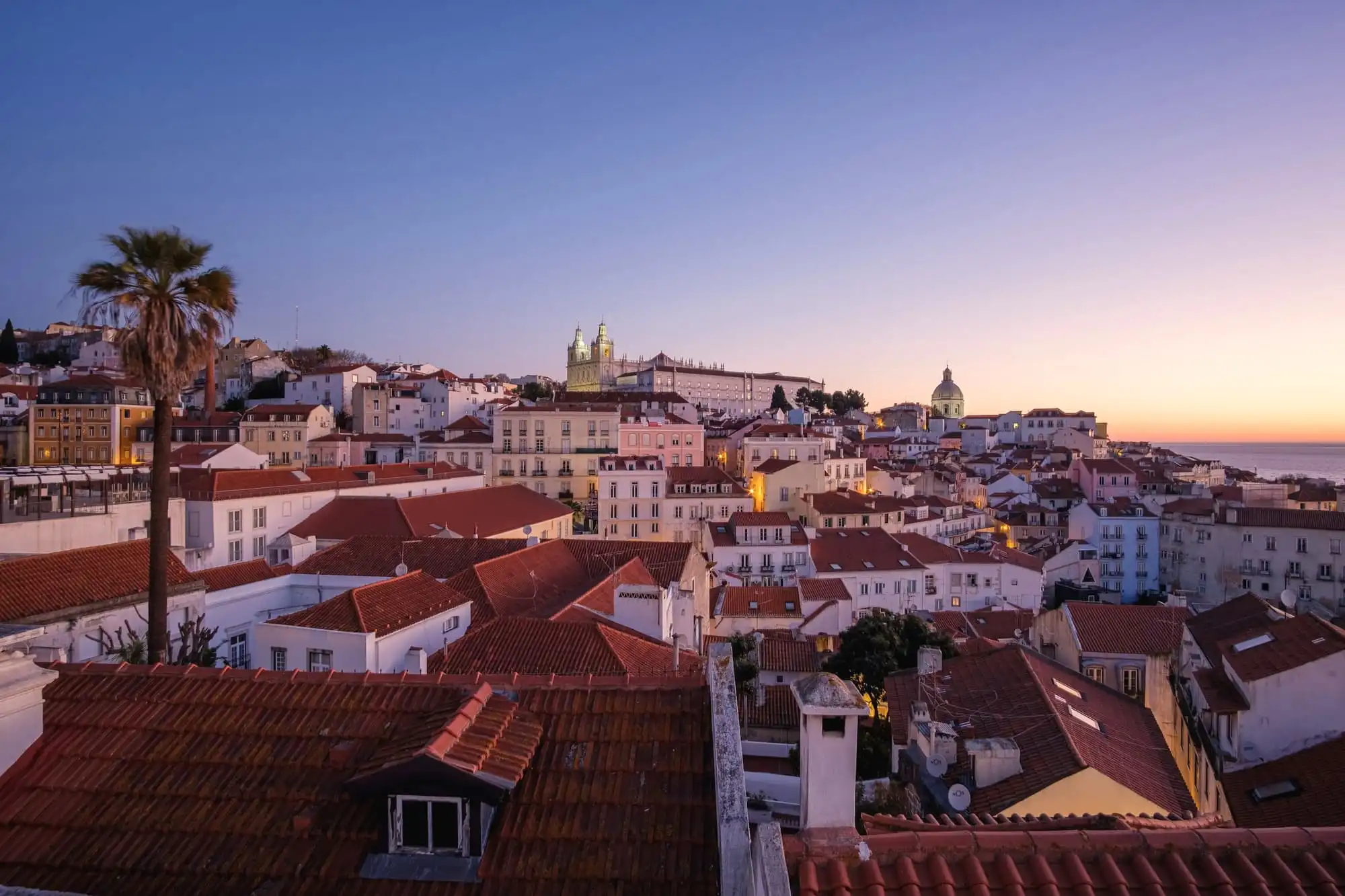 Alfama at dusk from a rooftop: terracotta roofs, white church domes and the river, under a violet sky turning orange at the horizon.
