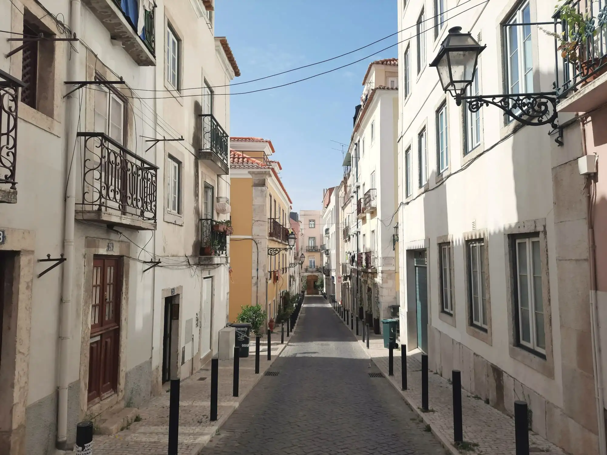 A narrow sunlit lane in Lisbon between tall white houses, with a lamp bracket on one wall and bollards along the pavement.