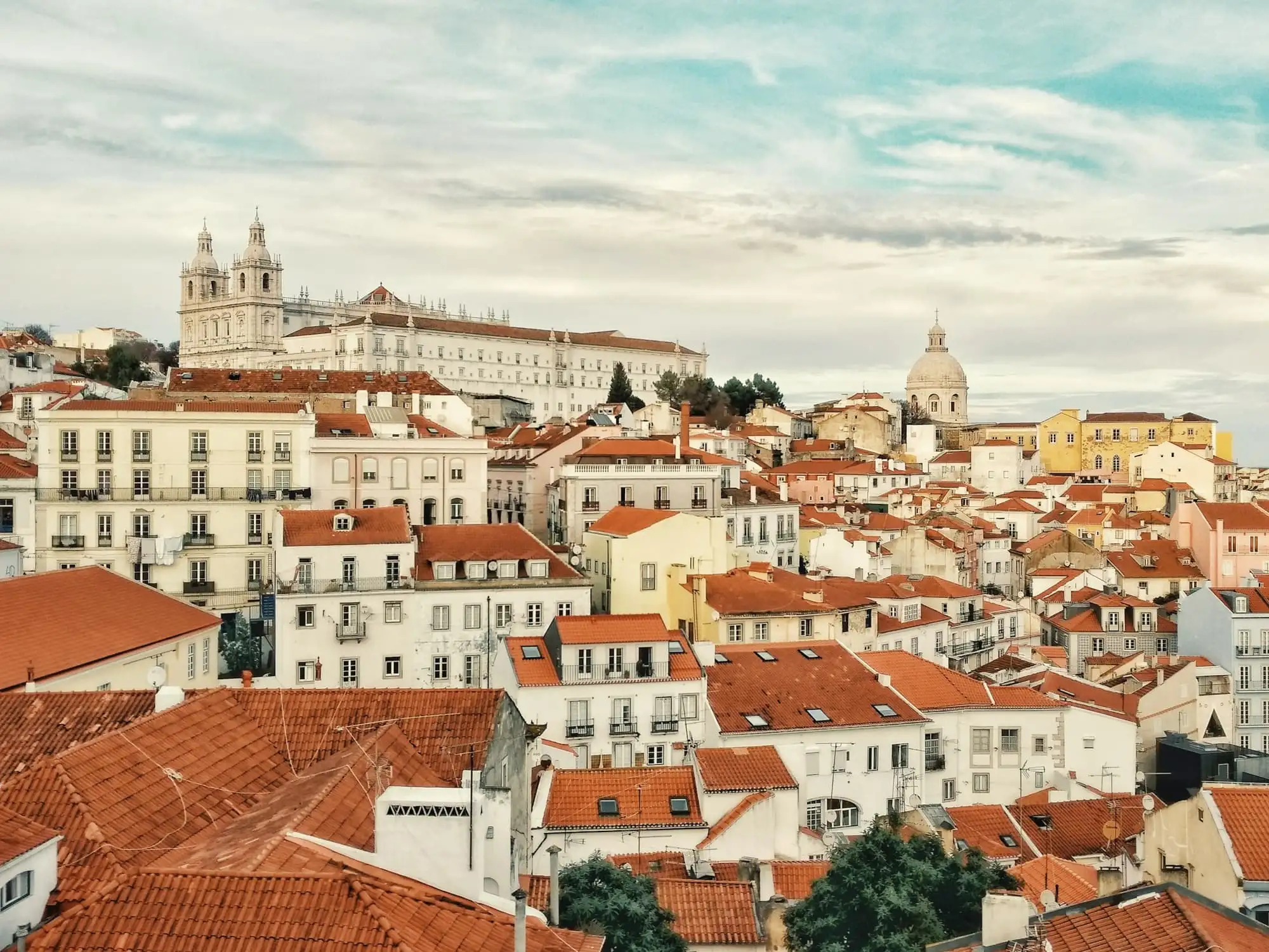 Red tiled roofs and white buildings of Alfama climbing a hill, with a monastery and a dome against a cloudy sky.