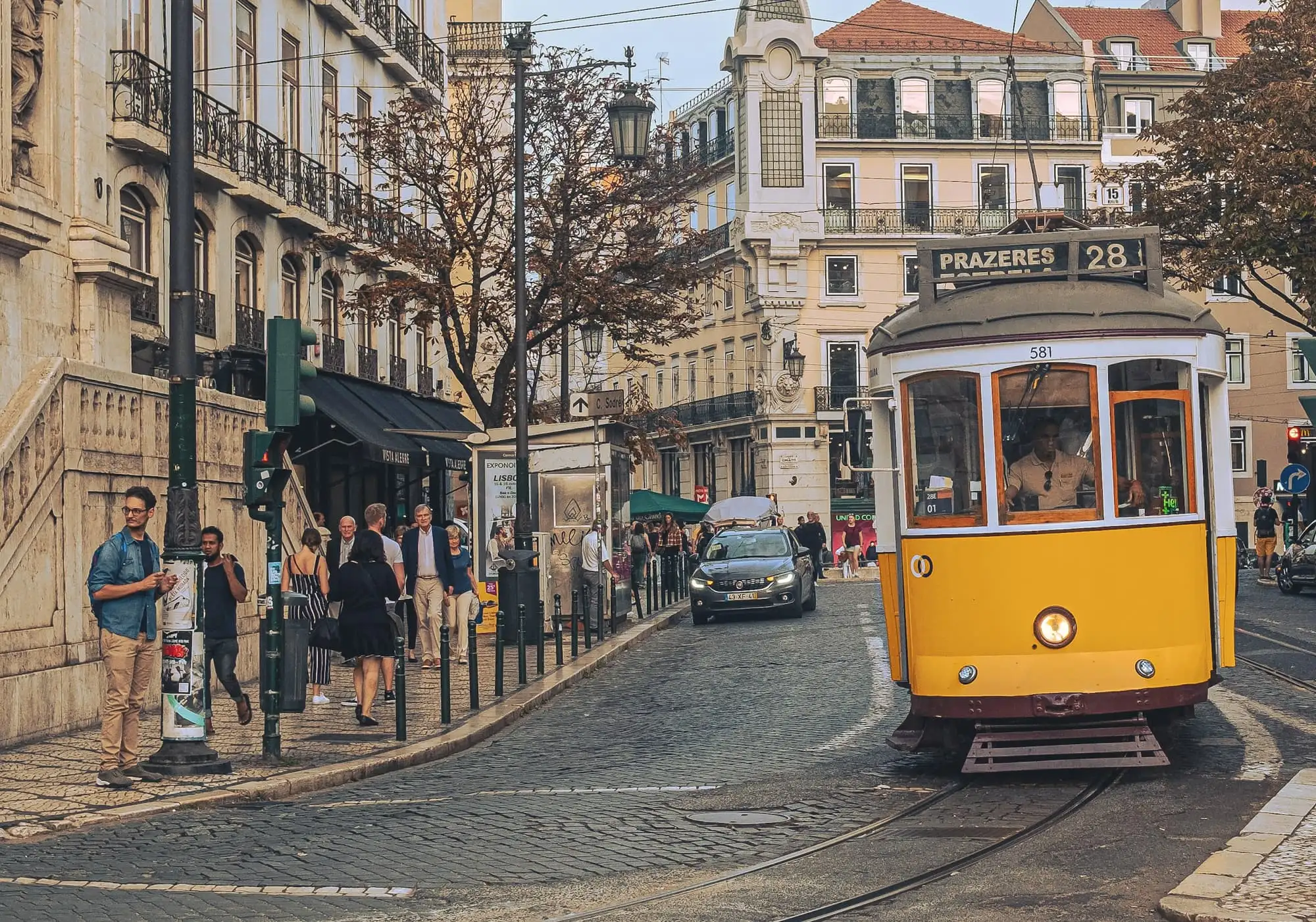 A yellow tram, number 28, on cobbled tracks in a street of tall pale buildings, with people on the pavement.