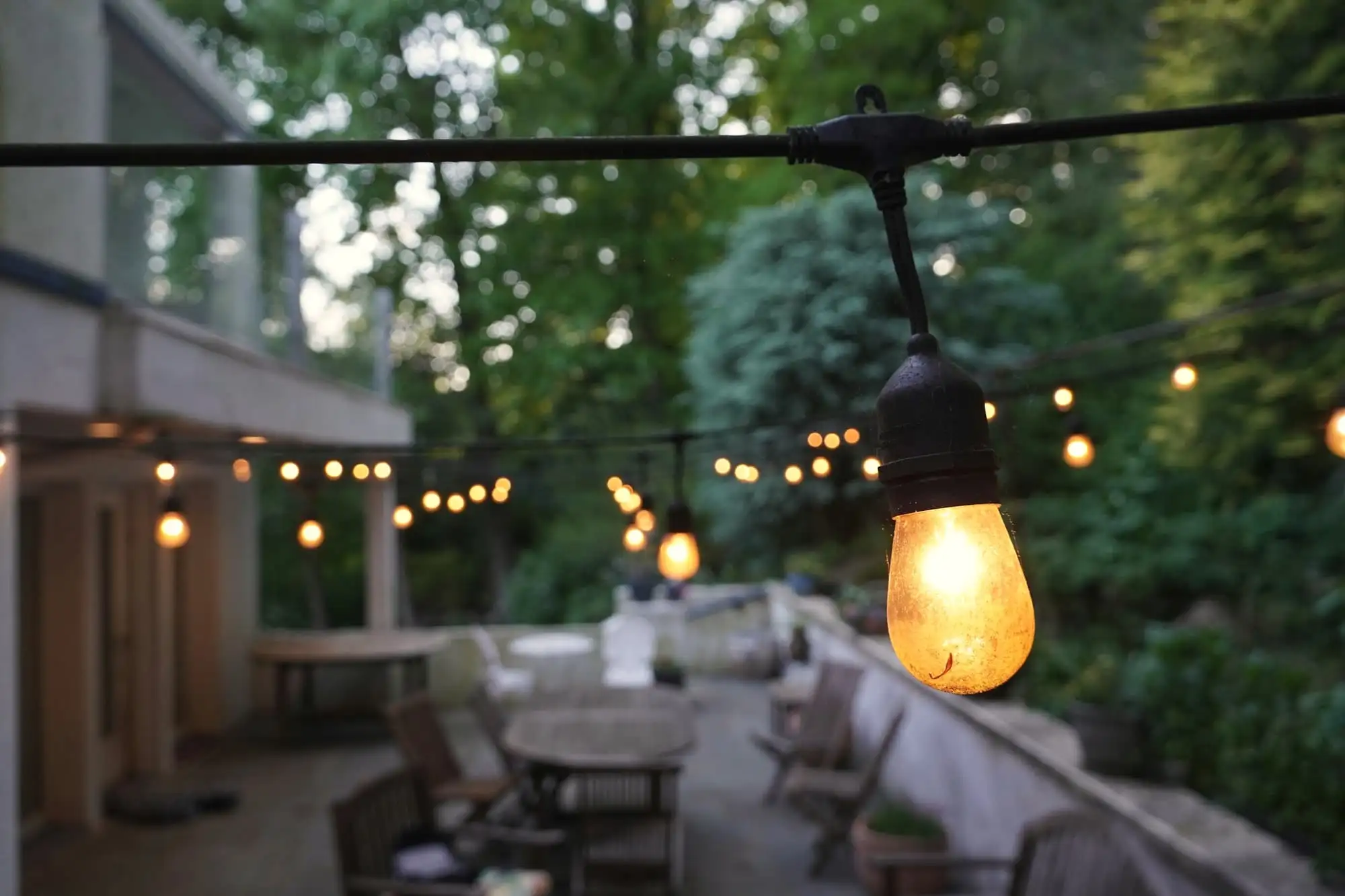 A string of warm bulbs over a quiet terrace with wooden tables and chairs, at dusk.